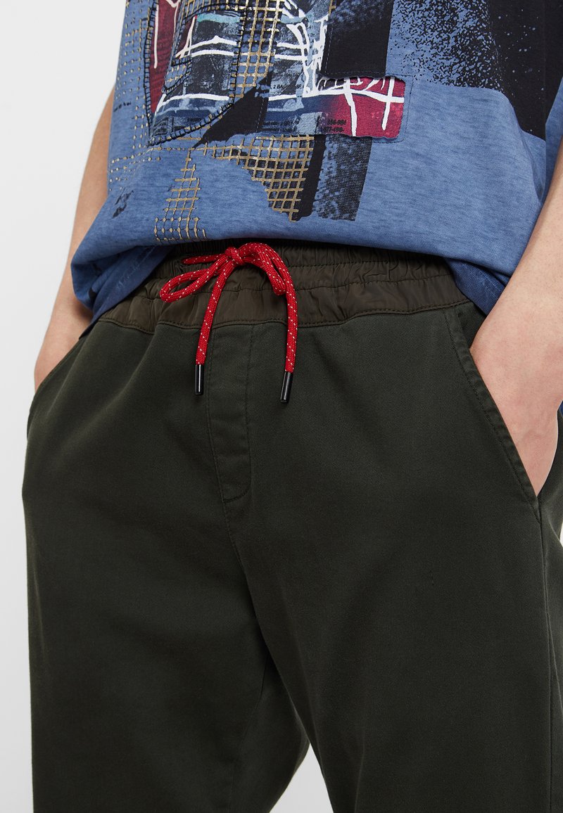 Olivgrüne Hosen mit einem Kordelzugbund, der eine rot- und silbergesprenkelte Schnur aufweist. Der Stoff wirkt glatt und hat seitliche Taschen.