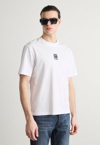 T-shirt en coton blanc à manches courtes et col rond, avec un petit logo gris imprimé sur la poitrine. Porté avec un jean en denim bleu.