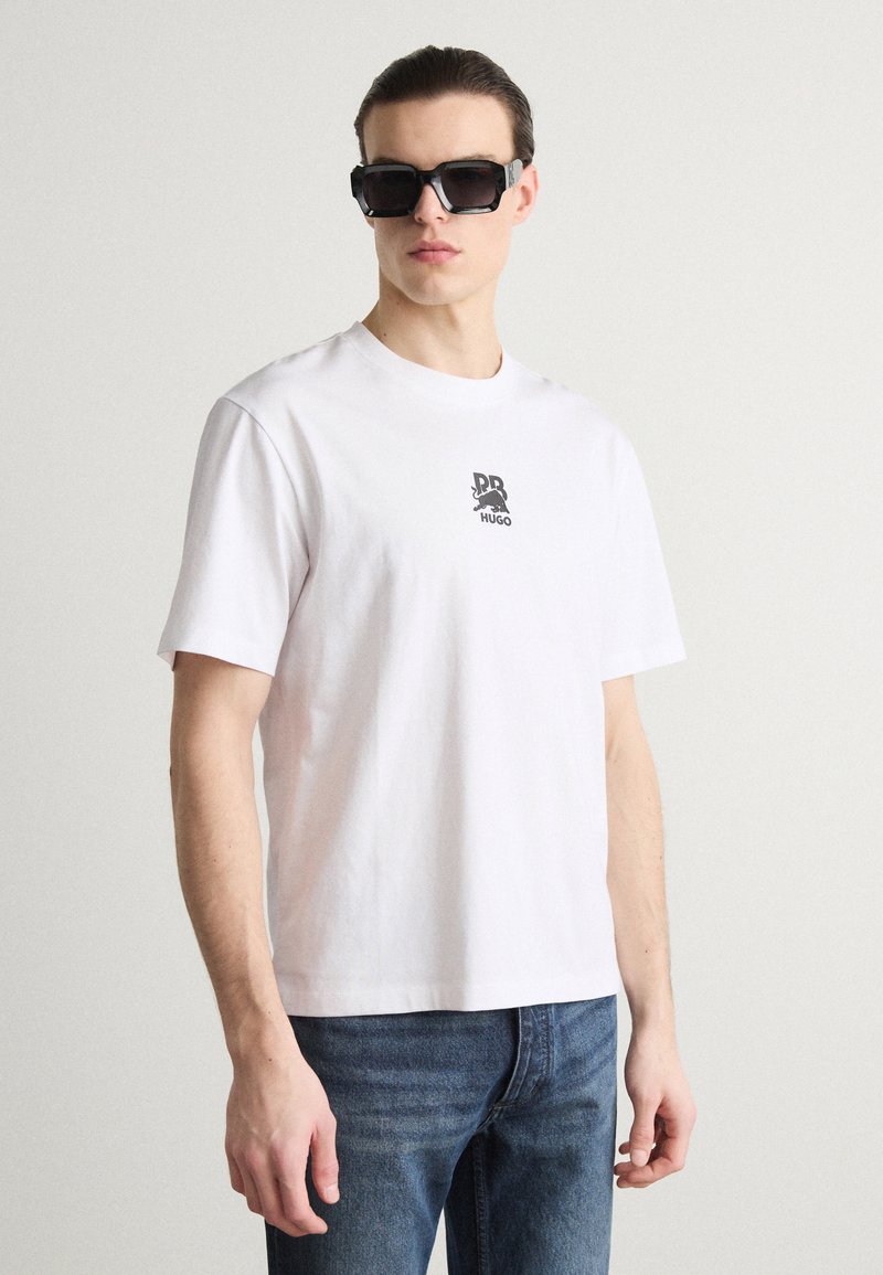 T-shirt en coton blanc à manches courtes et col rond, avec un petit logo gris imprimé sur la poitrine. Porté avec un jean en denim bleu.