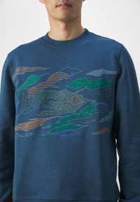 Homme portant un sweat-shirt bleu avec des motifs abstraits de montagnes et de nuages brodés en tons verts, marron et bleu.