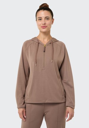 Frau steht frontal und trägt einen braunen Kapuzenpullover mit Reißverschluss vorne sowie eine passende Hose vor einem einfarbigen Hintergrund.