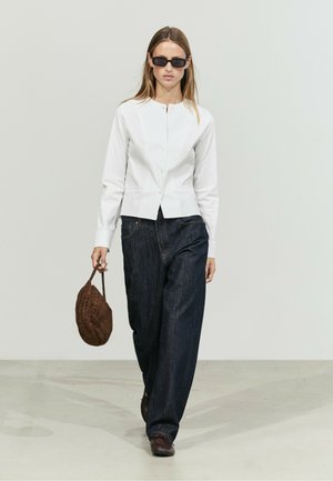 Femme portant des lunettes de soleil noires, un haut blanc boutonné, un jean large foncé, des chaussures marron et un sac à main marron texturé, marchant vers l'avant.