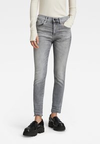 Jeans de mezclilla gris con una forma ajustada, diseño de cinco bolsillos y dobladillo deshilachado, combinados con mocasines negros de suela gruesa y una camiseta de manga larga blanca de canalé.
