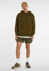 Felpa con cappuccio verde oliva con tasca frontale e logo, abbinata a pantaloncini verdi con motivo e sneakers verdi oliva, indossata con calzini bianchi.