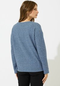 Donna in un maglione blu a maniche lunghe con piccoli punti bianchi sulle maniche, abbinato a pantaloni neri, in piedi contro uno sfondo bianco.