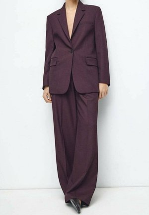 Person iført en dyb lilla skræddersyet jakkesæt med en enkeltknaps blazer og vidde bukser, stående mod en ensfarvet hvid baggrund.