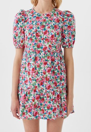 Kvinde iført en kort hvid kjole med puffede ærmer med et lyst pink, rødt, blåt og grønt blomstertryk.