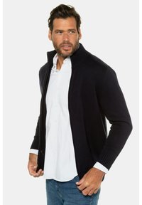 Mörkblå zip-up kofta med hög krage, ribbade muddar och texturerat stickat tyg, bärs över en vit skjorta och blå jeans.