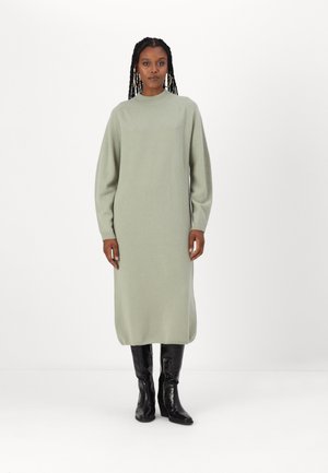 Knelengde, lys grønn strikket kjole med høy hals og lange ermer. Brukt med blanke svarte knelange støvler. Glatt tekstur, minimalistisk design.