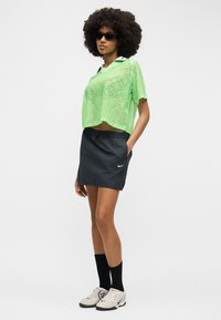 Γυναίκα με αφρο μαλλιά που φοράει μαύρα γυαλιά ηλίου, πράσινη διάφανη κοντομάνικη μπλούζα, μαύρη φούστα Nike, μαύρες κάλτσες και λευκά αθλητικά παπούτσια, όρθια.