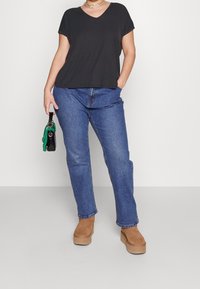 T-shirt nera a V abbinata a jeans in denim blu e stivali con zeppa color sabbia. Una piccola borsa nera con accenti verdi è tenuta nella mano sinistra.