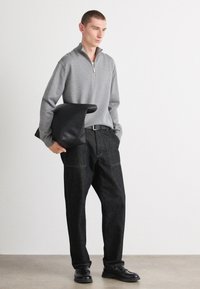 Grauer Reißverschluss-Pullover, schwarze Lederhandtasche, dunkle Jeans mit kontrastierenden Nähten und schwarze Schuhe. Einfaches Design, lockere Passform.