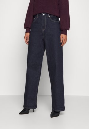 Person iført mørkeblå vidde jeans med høj talje, sorte spidse hæle og en vinrød langærmet sweater, der står foran en ensfarvet baggrund.