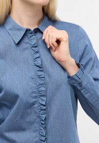 Blusa azzurra chiara con colletto, caratterizzata da una riga verticale di ruches sul davanti, polsini con bottoni e un tessuto testurizzato con sottili variazioni di colore.