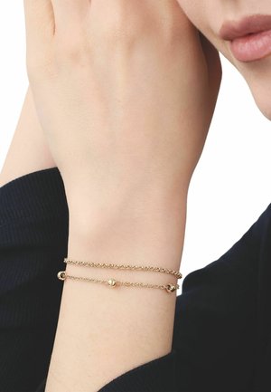 Muñeca con dos delicados brazaletes de cadena dorada, uno liso y otro con pequeñas cuentas redondas, sobre una camiseta negra de manga larga.