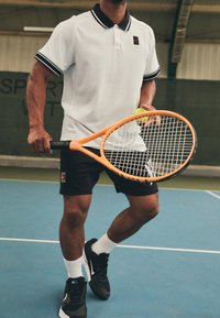 Polo bianca con colletto e dettagli neri sulle maniche, abbinata a pantaloni corti neri. Tieni in mano una racchetta da tennis arancione e una pallina, su un campo blu.