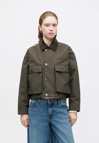 Olijfgroene cropped jas van gladde stof met een bruine corduroy kraag, voorzien van twee voorkruiken en zilveren drukknopen.