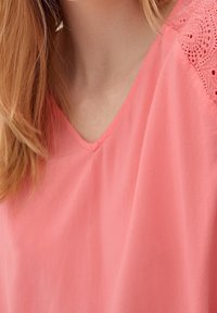 Blouse corail avec un décolleté en V et des accents en dentelle sur les épaules. Tissu doux avec une texture lisse, présentant une coupe décontractée.