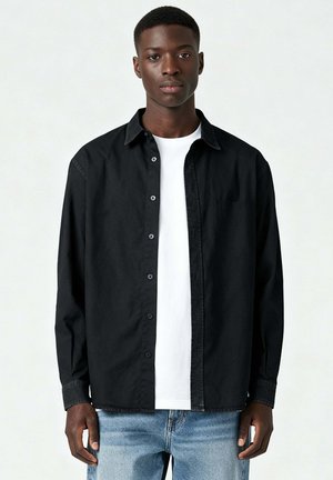 Jonge man die een zwart overhemd draagt over een wit T-shirt en lichtblauwe spijkerbroek, staand tegen een effen lichte achtergrond.