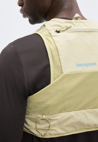 Άνδρας που φοράει μπεζ γιλέκο Patagonia πάνω από σκουρόχρωμο πουκάμισο, εμφανιζόμενος από τον ώμο μέχρι το μέσο της πλάτης, με έμφαση στο ύφασμα του γιλέκου και την μικρή τσέπη με φερμουάρ.