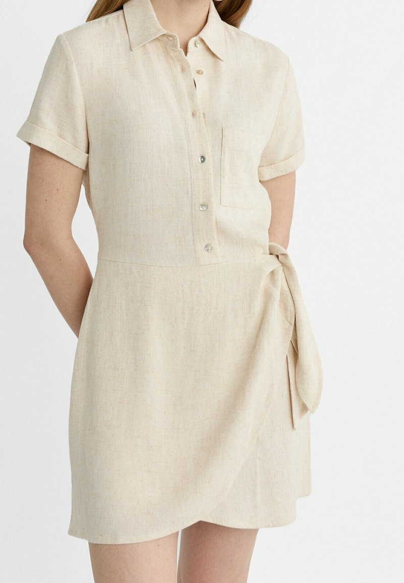 Vestito camicia in lino beige con colletto, maniche corte, bottoni frontali, taschino sul petto e cintura da annodare in vita. Tessuto morbido.