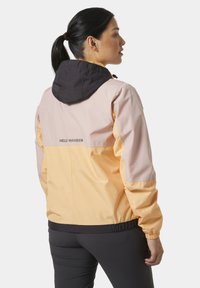 Chaqueta ligera a bloques de color en rosa pastel y melocotón con capucha negra. Presenta puños elásticos y un logo en la parte posterior.