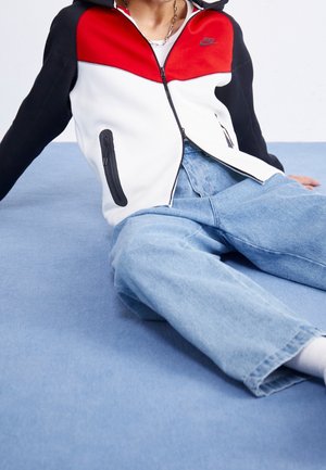 Person, die eine rot-weiß-schwarze Nike-Zipjacke und lockere hellblaue Jeans trägt, sitzt mit ausgestreckten Beinen auf einer blauen Fläche.