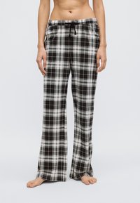 Kvinna som bär löst sittande pyjamasbyxor i svart, vitt och grått rutmönster med dragsko i midjan, stående barfota mot en enfärgad bakgrund.