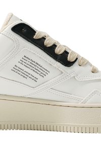 White leather sneaker with a textured rubber sole, black leather accents, and beige laces. Features a fabric label with brand information.