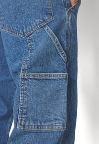 Pantaloni cargo din denim cu o nuanță de albastru ușor decolorată, având un buzunar lateral mare, cusături vizibile și o croială lejeră.