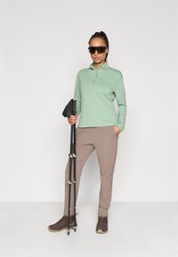 Ανοιχτό πράσινο φούτερ με φερμουάρ και μακριά μανίκια, καφέ ελαστικά παντελόνια, κρατώντας μπαστούνια πεζοπορίας, φορώντας σκούρα γυαλιά ηλίου και καφέ μποτάκια πεζοπορίας.