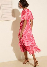 Robe midi rose à fleurs avec des manches courtes, une taille ajustée et un ourlet fluide. Le tissu transparent présente un motif tourbillonnant. Associée à des talons bruns.