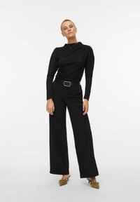 Črn dolg rokav s teksturirano tkanino in podrobnostmi na ramah, v kombinaciji s širokimi črnimi hlačami. Dopolnjeno s črnim pasom in zelenimi čevlji.