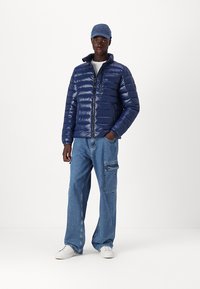 Navyblaue Pufferjacke mit glänzender Oberfläche, Reißverschluss und gerippten Bündchen, kombiniert mit hellblauen, weiten Jeans und weißen Turnschuhen.