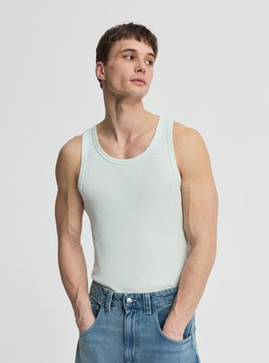 Giovane uomo che indossa una canotta a coste azzurro chiaro senza maniche e jeans blu, con le mani nelle tasche, guarda di lato su uno sfondo semplice.