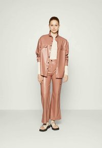 Conjunto de couro rosa com uma camisa de botão e calças de perna larga. A camisa tem dois bolsos frontais e um acabamento suave. Usado com sandálias estampadas.