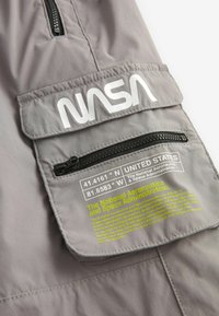 Sivý materiál s klopovou vreckom, na ktorom je biele písmeno "NASA". Obsahuje zipsové vrecko a potlač s údajmi a informáciami o súradniciach.
