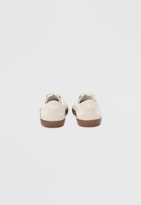 Tenisice s gornjim dijelom u kremasto-bijeloj boji izrađenim od teksturirane brušene kože i smeđim gumenim potplatom. Logotip „VEJA“ prikazan bijelom bojom na peti.