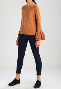 Bruine trui met wijde mouwen, gecombineerd met donkere skinny jeans en zwarte sneakers. Gladde textuur, losse pasvorm.