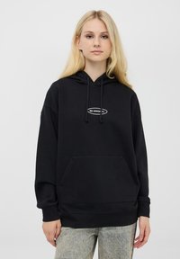 Czarna bluza z kapturem, wykonana z bawełny, z kieszenią kangurką, kapturem na sznurki oraz białym napisem "BĄDŹ ORYGINALNY" z przodu.