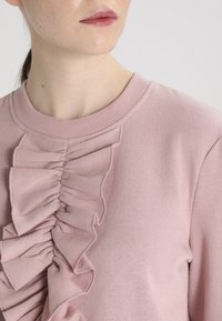 Sudadera rosa con detalle de volantes verticales en el frente, escote redondo y textura suave. Enfocada en un diseño casual y cómodo.