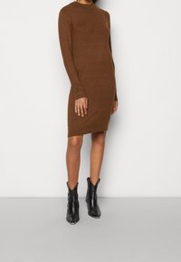 Abito maglione marrone a maniche lunghe, con polsini a costine e scollatura rotonda. Abbinato a stivaletti neri con punta.