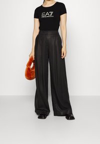 Camiseta negra de manga corta con logo de EA7, combinada con pantalones anchos negros. Sosteniendo un bolso de peluche naranja. Zapatos negros brillantes.