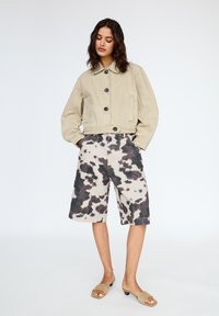 Giacca beige accorciata con grandi bottoni neri, abbinata a pantaloni corti fino al ginocchio con stampa a macchie di mucca nera e beige. Sandali con tacchi piccoli.