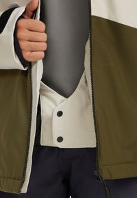 Veste présentant une partie supérieure crème, une partie inférieure vert olive, une fermeture éclair et des boutons-pression noirs ; inclut une doublure intérieure grise.