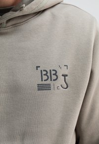 Lichtgrijze hoodie van een gestructureerde stof, met een bedrukt logo in donkergrijs dat "BB", een haak en horizontale lijnen omvat.