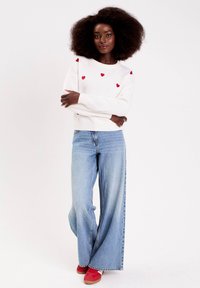 Femme avec des cheveux afro portant un pull blanc à cœurs rouges, un jean bleu à jambes larges et des baskets rouges, se tenant les bras croisés.