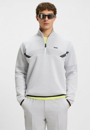 Ljusgrå zip-up tröja med en texturerad nätdesign, svarta accenter och ett kontrasterande neongult innerskikt. Bärs med grå byxor.