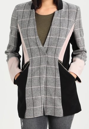 Vrouw draagt een op maat gemaakte blazer met een grijze geruite voorkant, zwarte zijkanten, lichtroze mouwen en ritszakken, gecombineerd met grijze jeans.