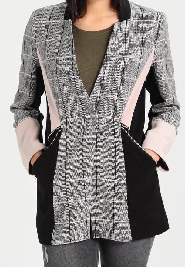 Femme portant un blazer sur mesure avec un devant à carreaux gris, des côtés noirs, des manches rose clair et des poches zippées, associé à un jean gris.